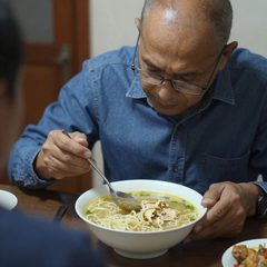 A man in his late sixties eating a bowl of chicken noodle soup at a table